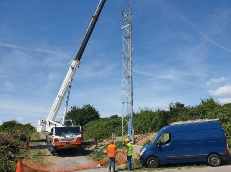Grue soulevant une antenne 5G avec deux ouvriers et une camionnette sur un chantier.