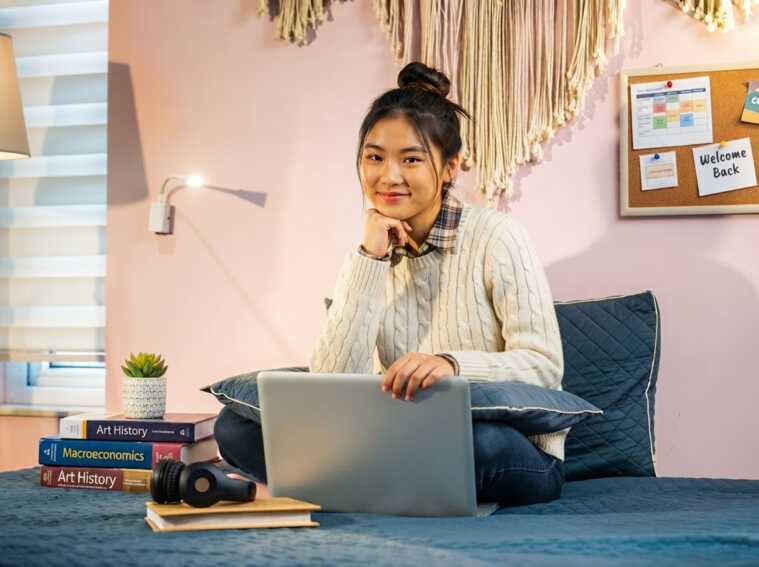 Jeune femme assise sur un lit avec un ordinateur portable, entourée de livres et d'un tableau d'affichage.