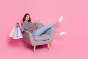 Femme dans fauteuil avec sas de courses et smartphone, heureuse de son iPhone 16 en soldes à 699€ chez Rakuten
