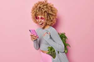 Femme coupe afro sur fond rose avec smartphone, très satisfaite de son forfait B&YOU de 100 Go à moins de 10€