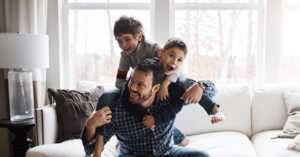 Papa avec 2 garçons sur le dos et smartphone à la main, joue et sourit, ravi de son nouveau forfait 5G B&YOU de 150Go à moins de 11€