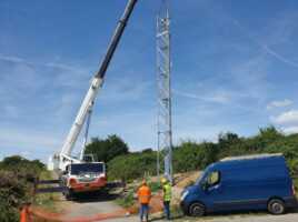 Grue soulevant une antenne 5G avec deux ouvriers et une camionnette sur un chantier.