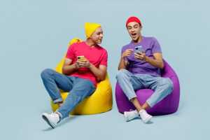 Deux garçons assis sur un pouf comparent leurs smartphones, contents de leurs forfaits YouPrice à moins de 10€ sur réseau Orange