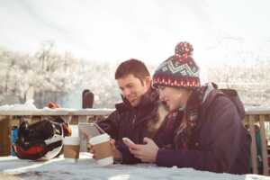 Couple de skieurs à bonnet en terrasse à la montagne, regardent leurs smartphones en souriant car forfaits de Lyca Mobile
