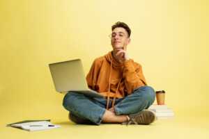 Jeune homme assis en tailleur avec PC portable sur fond jaune, gratuit dans le cadre des offres Box + High Tech de SFR