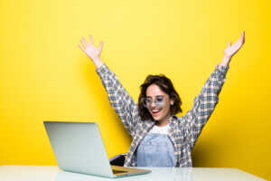 Femme devant PC portable sur fond jaune lève les bras de joie, contente de retrouver la SFR Fibre Power S