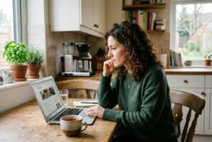 Femme de 30 ans aux cheveux bouclés lisant un article sur un ordinateur dans une cuisine.