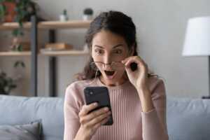 Femme avec smartphone leve ses lunettes et bouche ronde de surprise car elle paye trop cher son forfait mobile et peut trouver beaucoup mieux chez les MVNO