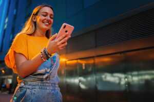 Femme en salopette et T-shirt Orange avec smartphone sourit, ravie de son forfait 40 Go à moins de 7€ chez YouPrice sur réseau Orange