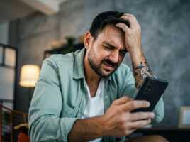 Un homme barbu avec un tatouage au bras tient un smartphone et se tient la tête avec une expression de douleur.