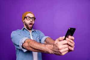 Homme sur fond violet avec smartphone à bout des bras, extrèmement surpris des 250€ de remise sur l'iPhone 16 chez Rakuten