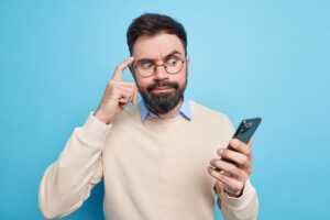 Homme barbu se gratte la tête devant son smartphone, se demandant si le reconditionné c'est bien