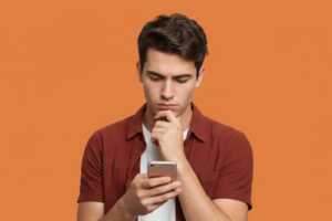 Jeune homme sur fond orange s'interroge devant son smartphone, pour savoir qui a le meilleur forfait avec 130Go entre Sosh, RED et B&YOU