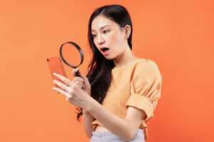Femme sur fond orange regarde son smartphone avec une loupe, car dur de trouver un modèle à 1€ en combo avec forfait mobile pas cher