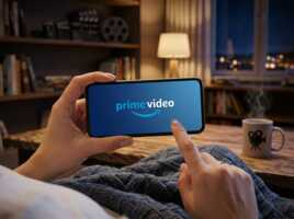 Mains tenant un smartphone affichant le logo Prime Video dans un salon avec une tasse et une bibliothèque.