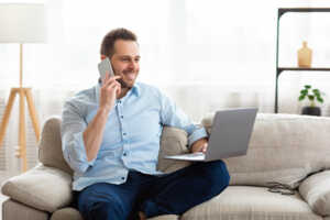 Homme dans son canapé, avec smartphone et PC portable, sourit car content du Wi-Fi 6E sur sa Box Internet