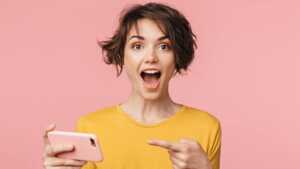 Femme sur fond rose montre son smartphone en souriant, ravie du forfait 5G de YouPrice avec déjà 130Go pour moins de 7€