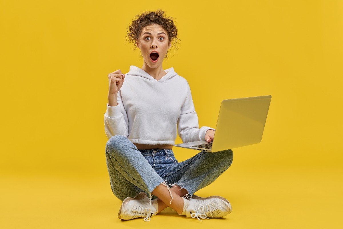 Femme sur fond jaune assise avec PC portable, bouche ronde de joie et surprise devant les accessoires gratuites et pratiques des Box Internet Premium