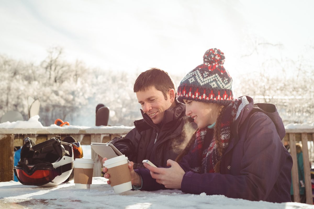 Couple de skieurs à bonnet en terrasse à la montagne, regardent leurs smartphones en souriant car forfaits de Lyca Mobile