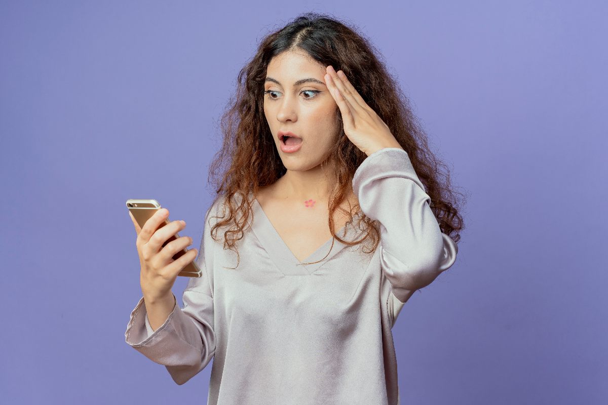 Femme sur fond violet se tient la tête et ouvre grands yeux et bouche en regardant son smartphone, choquée par l'excellent forfait de 80 Go en 5G chez Lyca Mobile