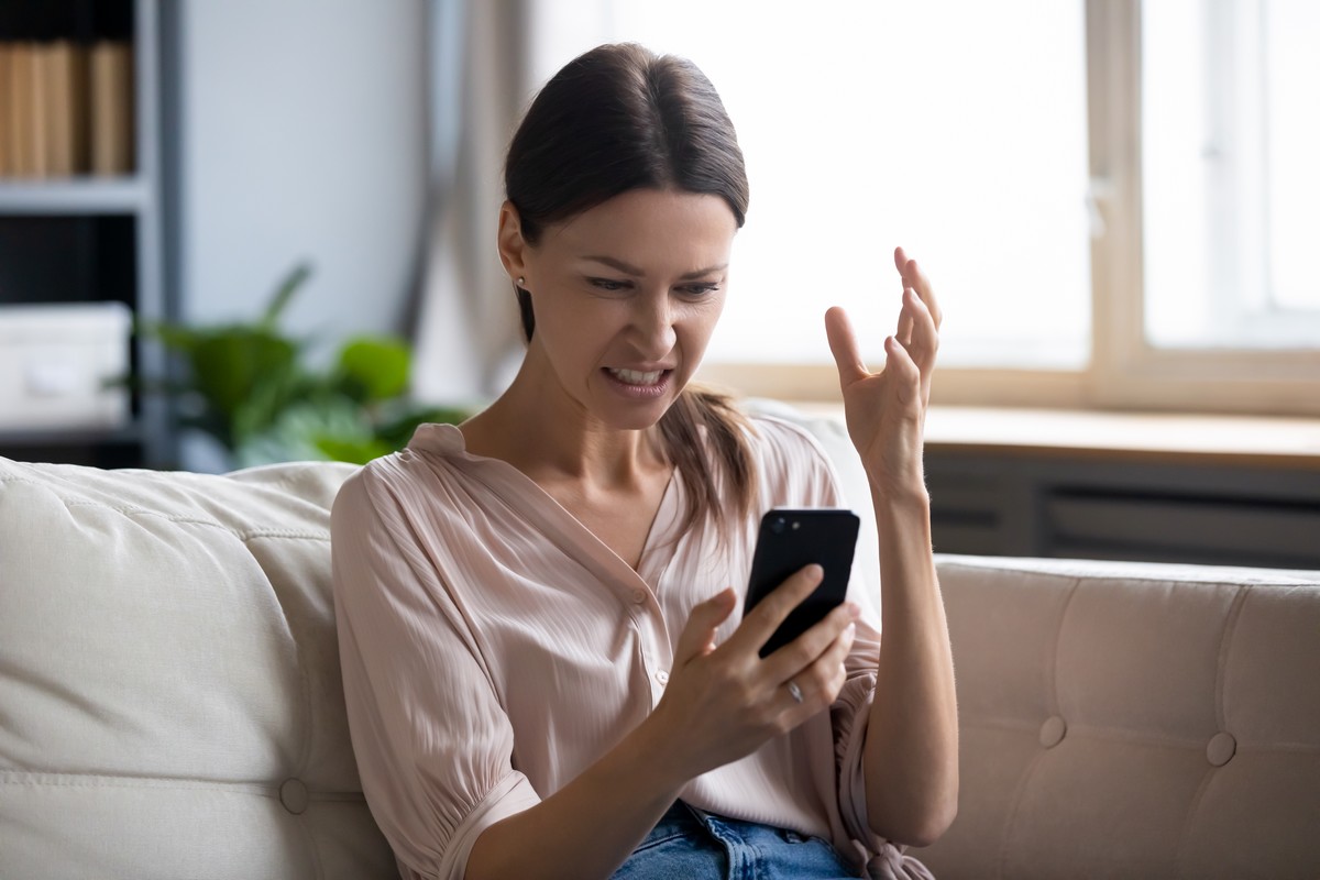 Une femme est énervée dans son canapé car son téléphone capte mal le réseau de son opérateur.