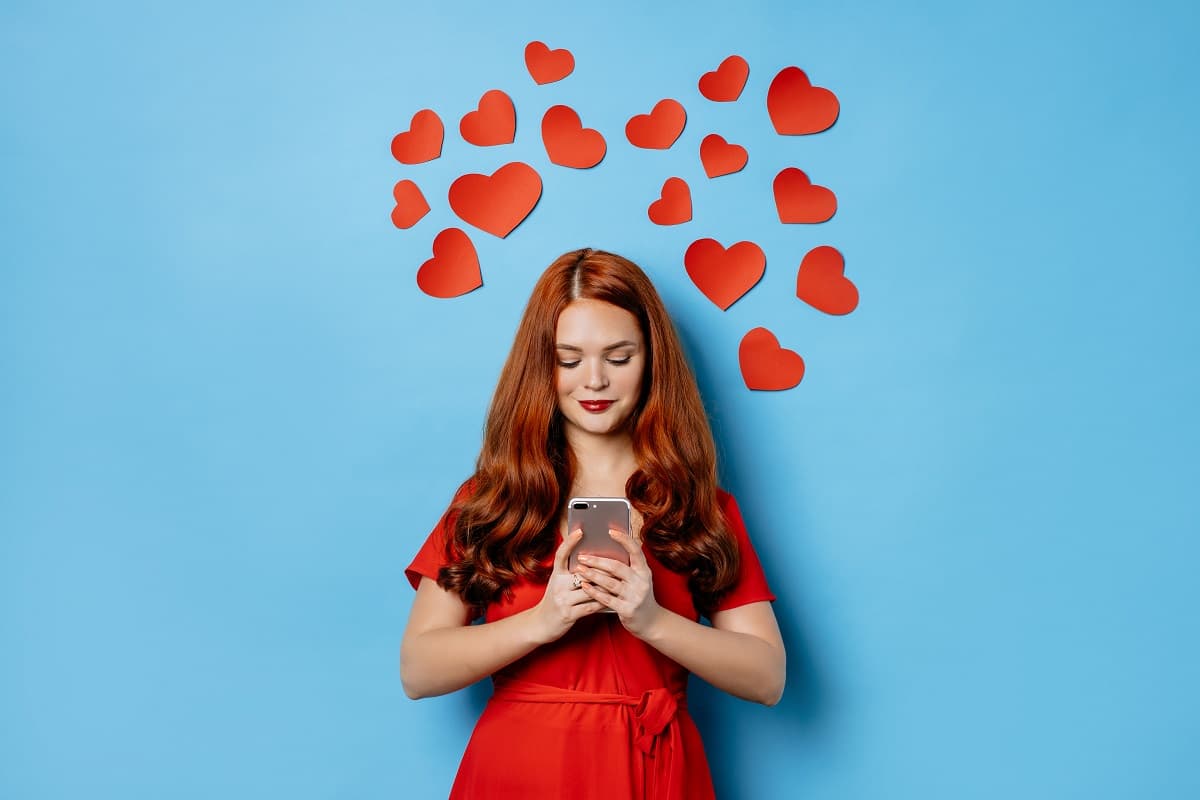 Femme en rouge avec des cœurs derrière elle et smartphone, ravie de son forfait promo de Saint Valentin de YouPrice
