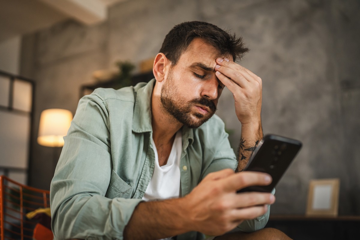 Homme souffle en regardant son smartphone, main sur la tête, stressé par les spams et appels frauduleux
