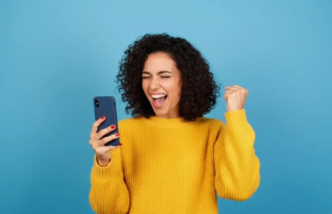 Femme avec smartphone sur fond bleu, serre le poing de joie et sourit, ravie de ces forfaits de 200 Go en 5G à moins de 10€