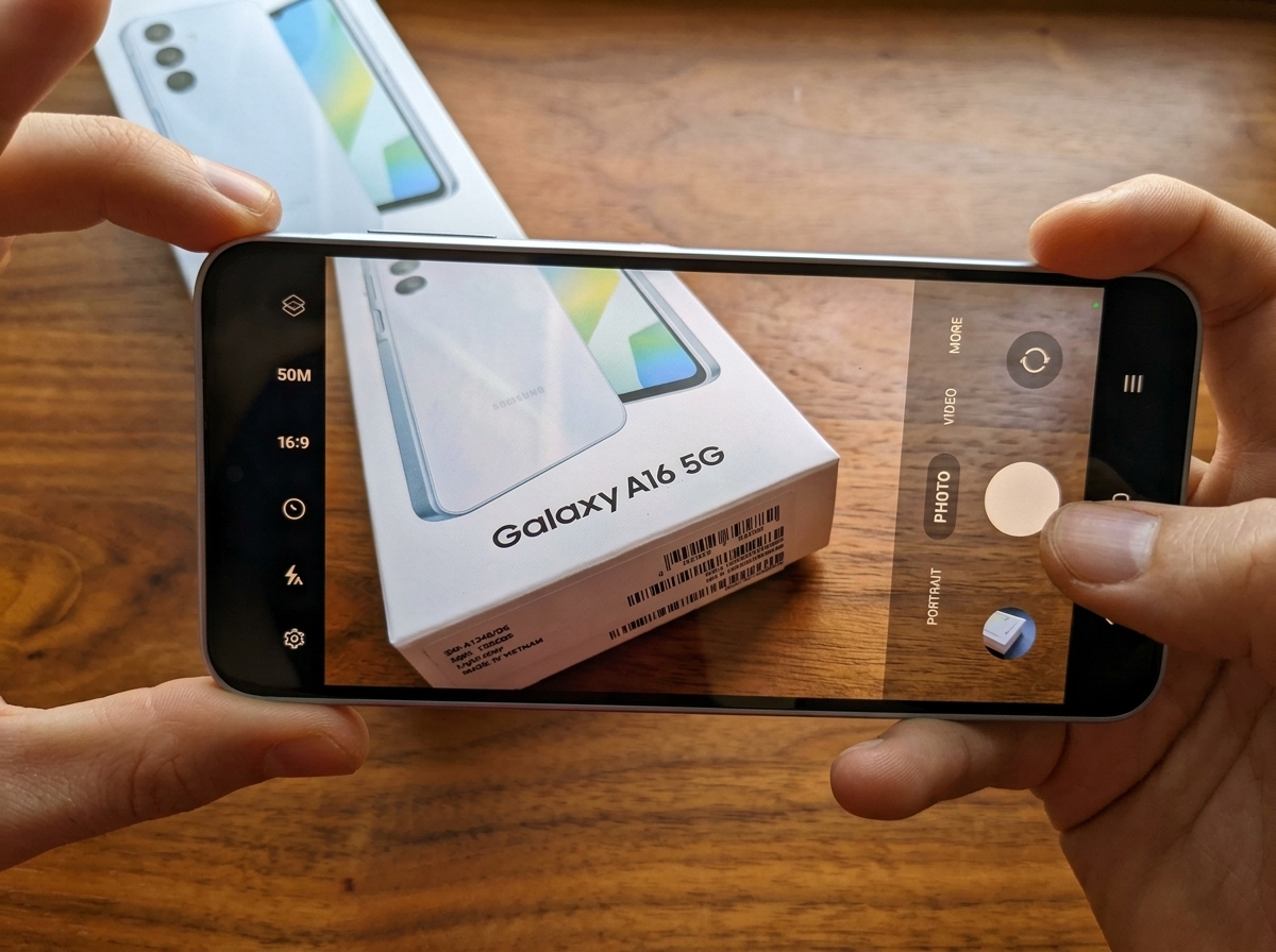 Mains tenant un smartphone Samsung Galaxy A16 5G photographiant sa boîte sur une surface en bois.