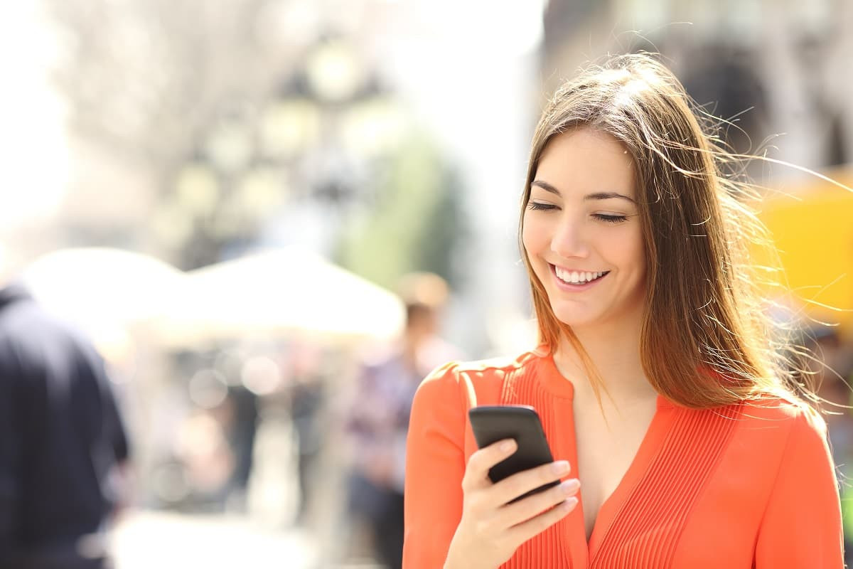 Femme en pull orange avec smartphone, ravie des code promo de Samsung