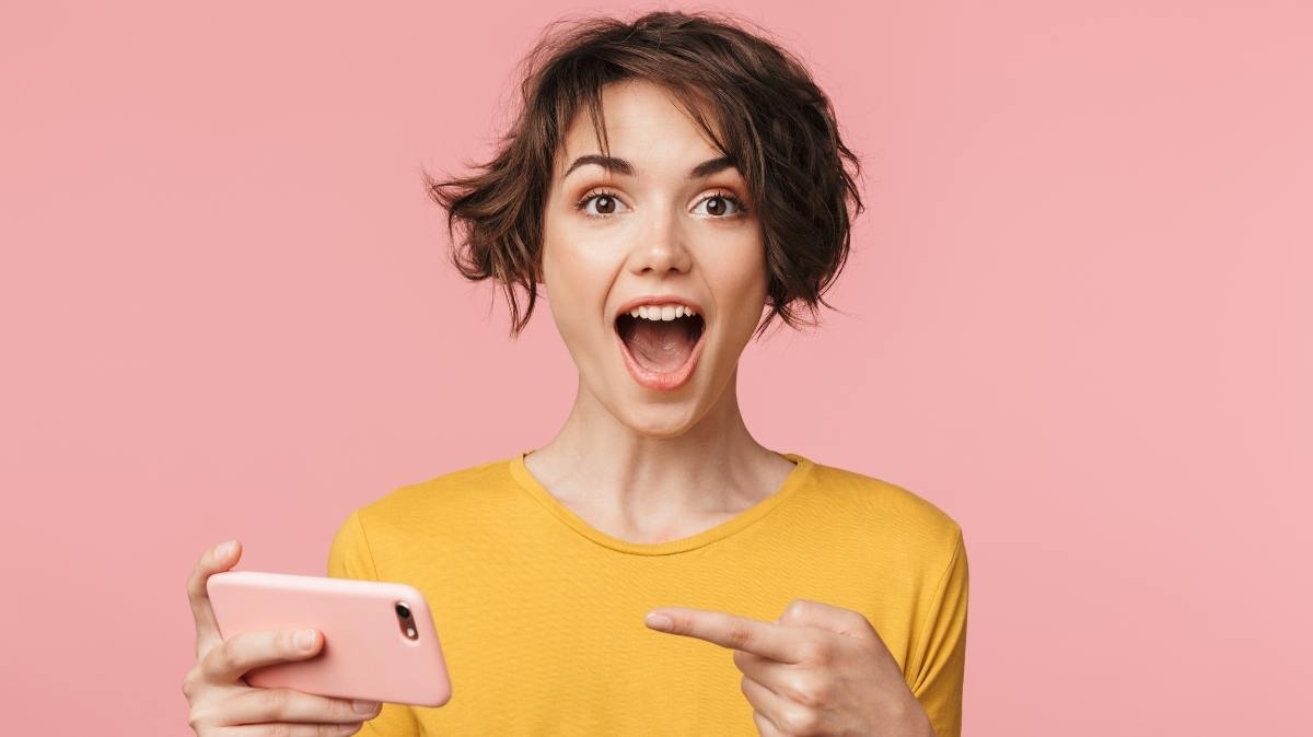 Femme sur fond rose montre son smartphone en souriant, ravie du forfait 5G de YouPrice avec déjà 130Go pour moins de 7€